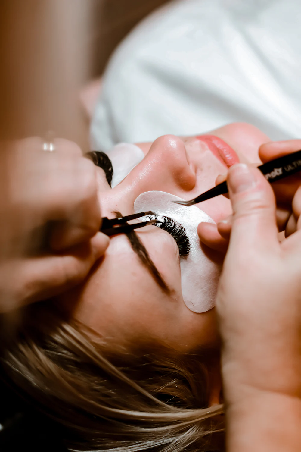 Profissional aplicando extensao de cilios com pinca em uma cliente deitada